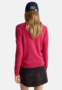 Maglione in maglia fucsia con orlo a coste, indossato sopra una gonna in velluto a coste nera. La modella indossa un berretto in denim scuro, mostrando i capelli lunghi.