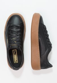 Sneakers in pelle nera con punta arrotondata e suola in gomma marrone testurizzata. Presentano lacci neri e logo del marchio dorato sulla linguetta.