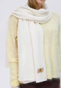 Sciarpa bianca realizzata in un morbido tessuto, con una texture liscia, disposta casualmente attorno a un maglione giallo chiaro, con un'etichetta di marca in legno visibile.