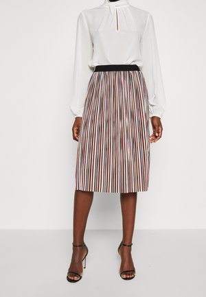Blouse blanche à manches longues avec un pli au niveau du col, associée à une jupe longueur genou ornée de rayures verticales dans des tons rouges, noirs et dorés.