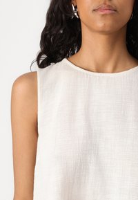Haut sans manches léger et texturé en tissu blanc cassé avec un col rond. Présente des variations subtiles de texture pour un intérêt visuel.