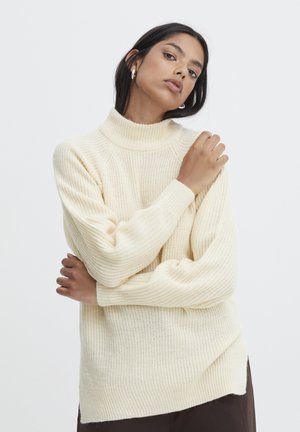 Pull en tricot couleur crème avec un col haut et côtelé. Présente des manches larges, des motifs texturés et une coupe décontractée. L'ourlet est légèrement asymétrique.