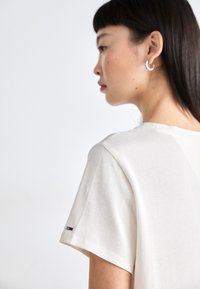 Camiseta de rayas crema con escote redondeado y mangas cortas. Presenta un pequeño logotipo en la manga izquierda. Textura suave.