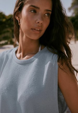 Junge Frau mit langen dunklen Haaren, die ein hellblaues ärmelloses Oberteil und goldene Creolen trägt, schaut im sanften Tageslicht im Freien weg.