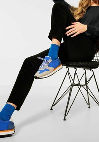 Niebiesko-szare buty sportowe z białymi akcentami i pomarańczową podeszwą, w połączeniu z niebieskimi skarpetami. Czarne spodnie i czarne krzesło w minimalistycznym otoczeniu.