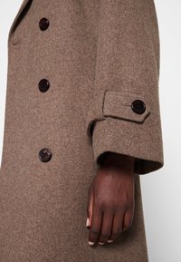 Brun uldfrakke med en struktureret design, der har mørke knapper og en manchet med en flap. Nærbillede af ærmet og hånden.