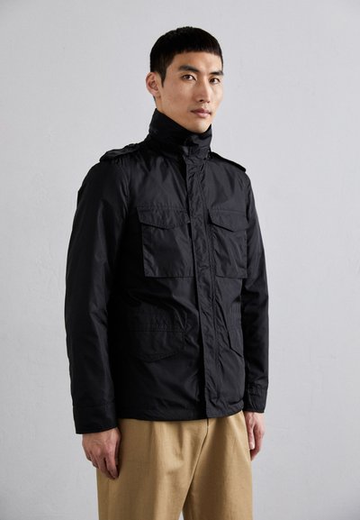 Veste noire imperméable avec un col montant, plusieurs poches avant et une fermeture éclair, assortie à un pantalon beige, se tenant contre un fond neutre.