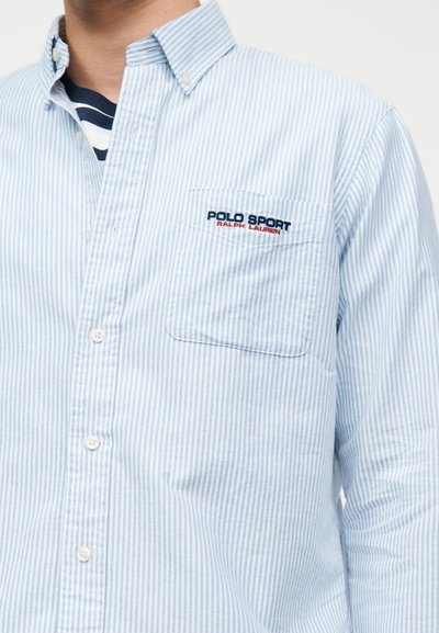 Ανοιχτό μπλε και λευκό ριγέ πουκάμισο με κουμπιά, με τσέπη στο στήθος κεντημένη με τη φράση "Polo Sport Ralph Lauren" σε ναυτικό μπλε και κόκκινα γράμματα.