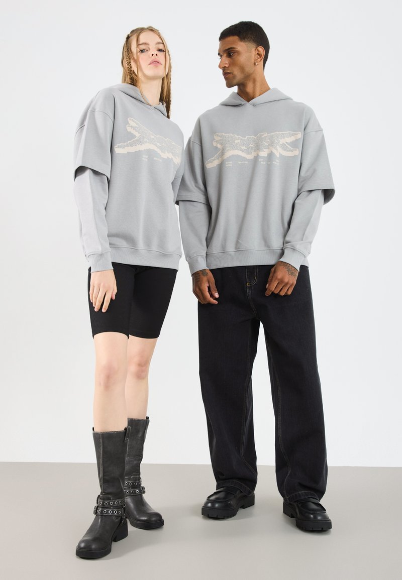 Sudaderas grises con un diseño gráfico, combinadas con pantalones cortos de ciclismo negros y botas negras hasta la rodilla con correas. Diseño simple y de corte relajado.