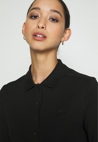 Polo shirt nera con colletto classico, tessuto strutturato e patta dei bottoni. Logo ricamato di colore abbinato sul petto.