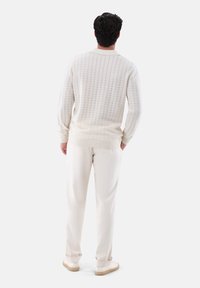 Mann trägt einen strukturierten weißen Pullover mit langen Ärmeln, cremefarbene Hose und weiße Sneakers, steht mit hinter dem Rücken verschränkten Händen und schaut weg.