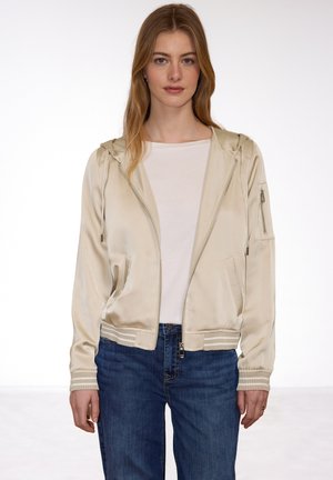 Young woman with long blonde hair wearing a cream satin zip-up hoodie, white shirt, and blue jeans standing against a white background.