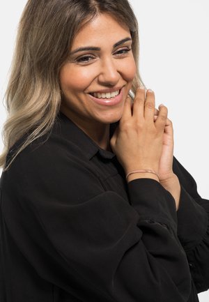 Blouse noire à manches longues avec des manches bouffantes ; modèle portant un bracelet métallique léger ; peau douce et lisse avec un maquillage neutre ; fond clair.
