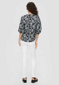 Zwart-witte blouse met bloemmotief en ballonmouwen, gecombineerd met witte broek en zwarte platform sandalen. Effen achtergrond.