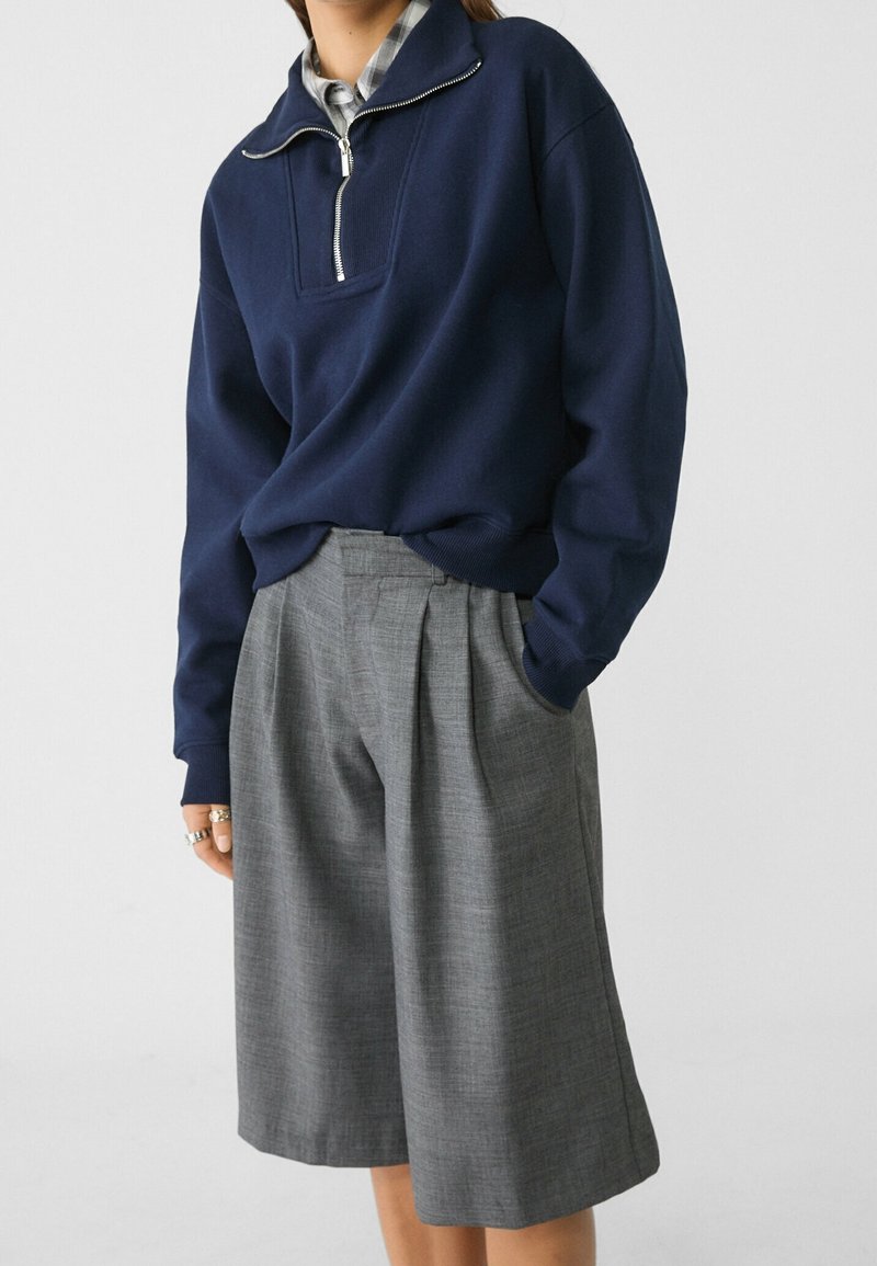 Personne portant un sweat-shirt bleu marine à fermeture éclair quart-de-course superposé à une chemise à carreaux, associé à un short large gris plissé taille haute.