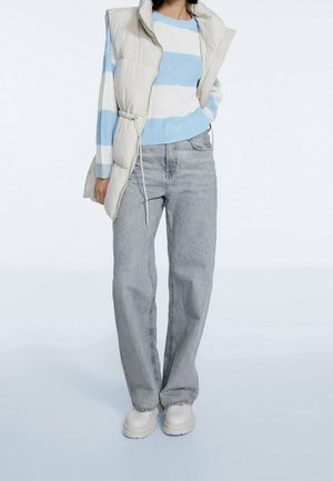 Personne portant un pull tricoté rayé bleu et blanc, un gilet matelassé sans manches beige, un jean gris clair à jambes droites, et des chaussures blanches épaisses.