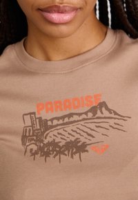 Un t-shirt marron présente un design graphique avec "PARADISE" en orange au-dessus d'une plage bordée de palmiers et d'un paysage montagneux en marron foncé.
