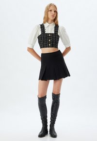 Zwart tweed crop top met rafelige randen en gouden knopen, gecombineerd met een zwarte geplooide mini-jurken. Kniehoge zwarte leren laarzen maken de look compleet.