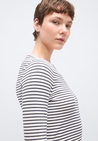 Fehér és tengerészkék csíkos hosszú ujjú felső, testhezálló szabással és puha anyagból, kerek nyakkivágással és diszkrét oldalsó varrási részletekkel.