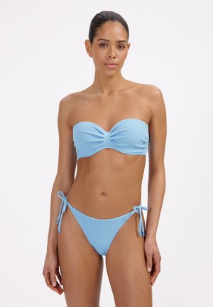 Kobieta ubrana w jasnozielony teksturowany bikini bez ramiączek z wiązanymi po bokach spodenkami, stojąca na tle białego tła.