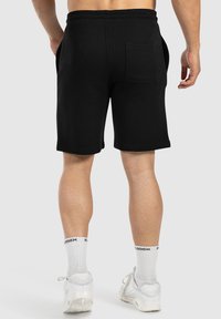 Mann in schwarzen knielangen Shorts, weißen Kniestrümpfen mit schwarzem Text und weißen Sneakers, von hinten vor einem schlichten Hintergrund gezeigt.