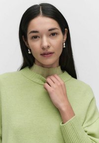 Jasnozielony sweter dziany z prążkowanym kołnierzem i teksturowanymi rękawami; model delikatnie dotyka kołnierza, nosząc srebrne kolczyki.