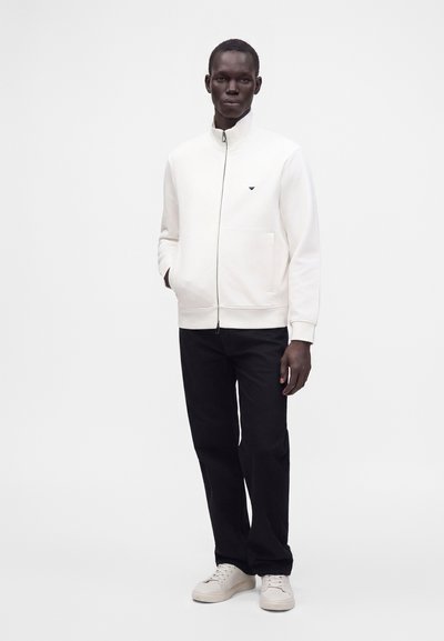 Homme portant une veste blanche zippée, un pantalon noir et des baskets blanches, debout devant un fond blanc uni avec une main dans la poche.