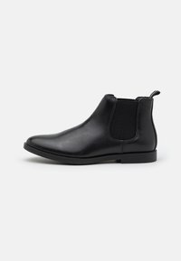 Botín Chelsea de cuero negro con paneles elásticos a los lados, tacón bajo, punta redondeada y un asa en la parte trasera. Textura suave y diseño elegante.