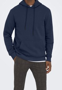 Felpa blu navy realizzata in tessuto morbido, con tasca frontale, lacci sul cappuccio e polsini a costine, abbinata a pantaloni a quadri marroni.