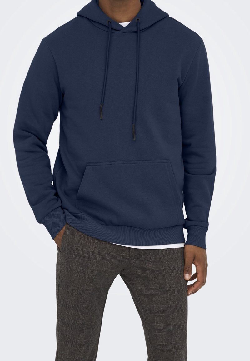 Felpa blu navy realizzata in tessuto morbido, con tasca frontale, lacci sul cappuccio e polsini a costine, abbinata a pantaloni a quadri marroni.