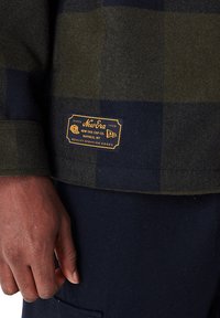 Olijfgroen en marineblauwe geruite wollen shirt met een gestructureerde afwerking. Bevat een gouden New Era-label nabij de zoom, met contrasterende stiksels.