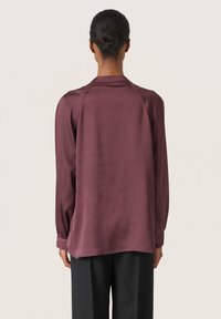 Maroonblus med ett flödande mönster, som har långa ärmar och en plisserad textur. Skräddarsydd passform med en subtil lyster, bärs tillsammans med svarta byxor.