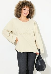 Sudadera oversized beige con texto "Libertad" en acabado brillante. Tejido suave, mangas anchas y aberturas en el dobladillo. Combinada con jeans oscuros.