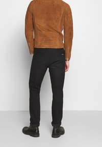 Giacca in suede marrone con schiena texture e dettagli a costine, abbinata a jeans neri e stivaletti neri, vista da dietro.