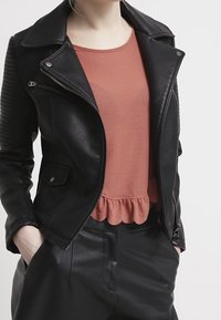 Chaqueta de cuero sintético negra con cremalleras, bolsillos con solapa y acentos acanalados en los hombros, combinada con una blusa de manga corta color óxido y pantalones negros.