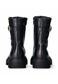 Stivali in pelle nera fino al polpaccio con suole spesse e scanalate e cinghie con fibbie laterali, mostrati da dietro su uno sfondo bianco.