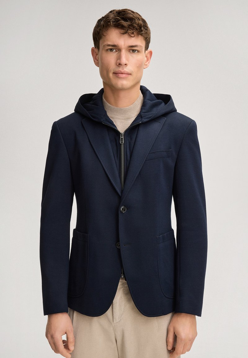 Blazer bleu marine avec capuche, en tissu structuré. Comprend deux poches avant et une fermeture à un bouton. Porté sur un haut beige clair.