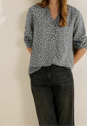 Kobieta w szarej bluzce z długim rękawem z białymi kwadratowymi wzorami i czarnych dżinsach, stojąca z rękami za plecami na tle gładkiej ściany.