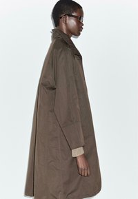Cappotto trench oversize marrone con colletto alto e tessuto texturizzato. Presenta una vestibilità comoda e maniche lunghe. Indossato sopra un maglione beige.