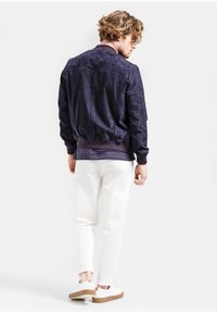 Námornícka zamšová bomber bunda s rebrovanými manžetami a spodným lemom, nosená s bielymi nohavicami a béžovými topánkami, s uvoľneným strihom a textúrovaným materiálom.