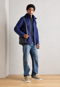 Veste imperméable navy avec col haut, portée par-dessus une chemise noire. Jeans bleu clair et baskets beiges. Sac en bandoulière noir. Texture lisse.