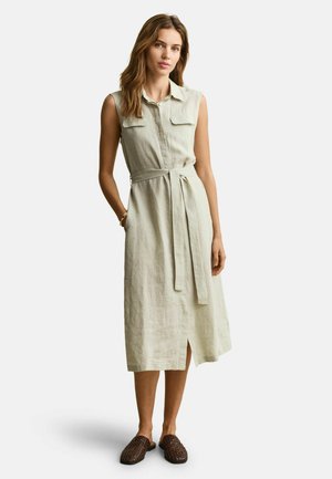 Vrouw die een mouwloze beige midi-jurk met ceintuur en zakken aan de voorkant draagt, staand met haar handen gedeeltelijk in de zakken en bruine geweven instapschoenen aan.