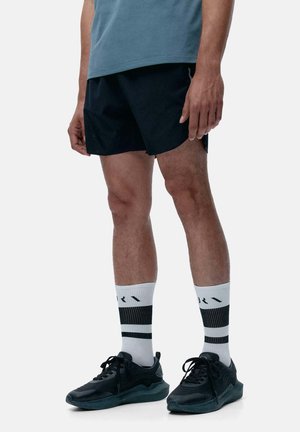 Shorts de sport noirs, t-shirt gris, chaussettes blanches avec des rayures noires et baskets noires. Jambes visibles avec des poils clairs et un arrière-plan neutre.
