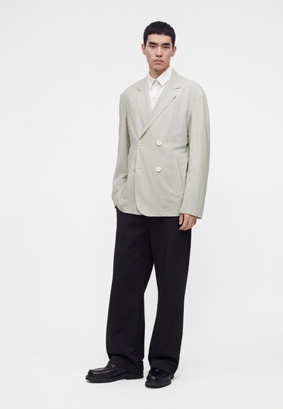 Jeune homme portant un blazer croisé gris clair, une chemise blanche, un pantalon noir à jambes larges et des chaussures noires, debout devant un fond uni.