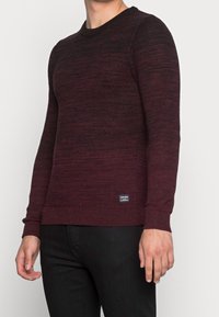 Burgundy stickad tröja med ett strukturerat mönster, rund halsringning och ribbade ärmslut och nederkant. Har en liten märkesetikett på botten.