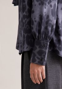 Blusa a stampa animale nera e grigia con una vestibilità comoda, maniche lunghe, polsini con bottoni e una texture di stoffa morbida. Indossata sopra un capo grigio.