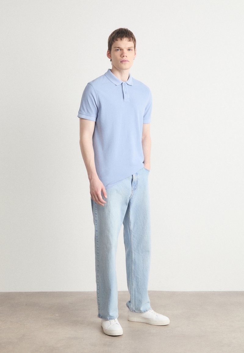 Chemise polo bleu clair en coton, à manches courtes avec des poignets côtelés, associée à un jean en denim clair et des baskets blanches. Fond neutre.