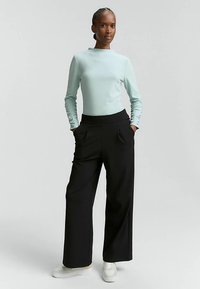 Hellblaues Langarmshirt und schwarze Hosen mit weitem Bein. Die Hosen haben seitliche Taschen; weiße Sneakers vervollständigen das Outfit.