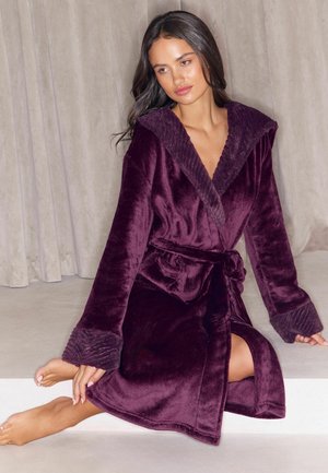 Mujer con cabello largo y oscuro, vestida con una bata mullida de color púrpura oscuro, sentada con una rodilla doblada y mirando hacia un lado frente a un fondo de cortina beige.