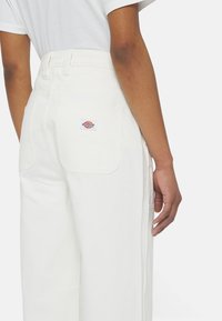 Vita vida byxor i bred modell gjorda av slitstark tyg, med en bakficka och en märkesetikett med blå och röda inslag.